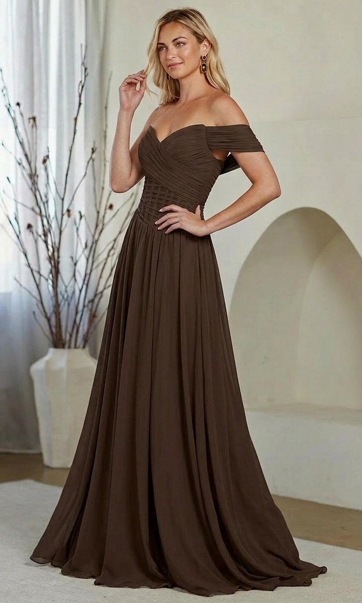 A woman stands indoors wearing the Amelia Couture 9097, a long, off-the-shoulder prom dress featuring a fitted bodice and flowing A-line skirt in rich brown.